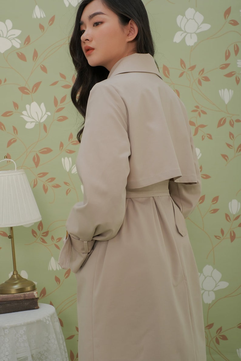 K&ouml;nnte beinhalten: Ein beiger Trenchcoat mit G&uuml;rtel und Kragen. Der Mantel ist aus glattem Stoff und hat ein klassisches Design.