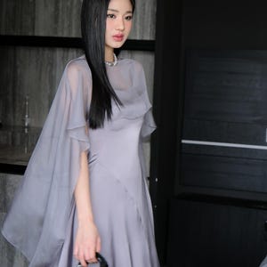 Pode incluir: Uma mulher em um vestido cinza com uma capa transparente combinando e uma pequena bolsa preta. O vestido tem uma saia fluida e um corpete ajustado. A capa cai sobre os ombros, criando uma silhueta suave e elegante. A bolsa &eacute; pequena e brilhante.