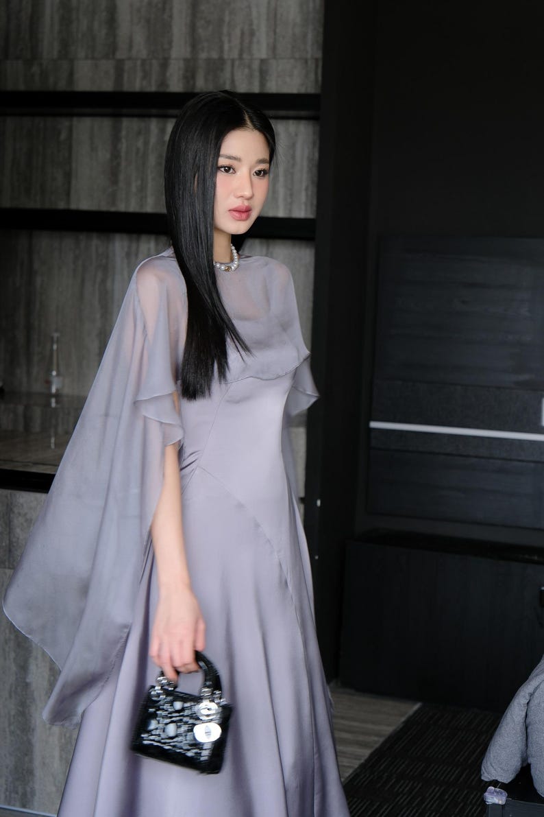 Pode incluir: Uma mulher em um vestido cinza com uma capa transparente combinando e uma pequena bolsa preta. O vestido tem uma saia fluida e um corpete ajustado. A capa cai sobre os ombros, criando uma silhueta suave e elegante. A bolsa &eacute; pequena e brilhante.