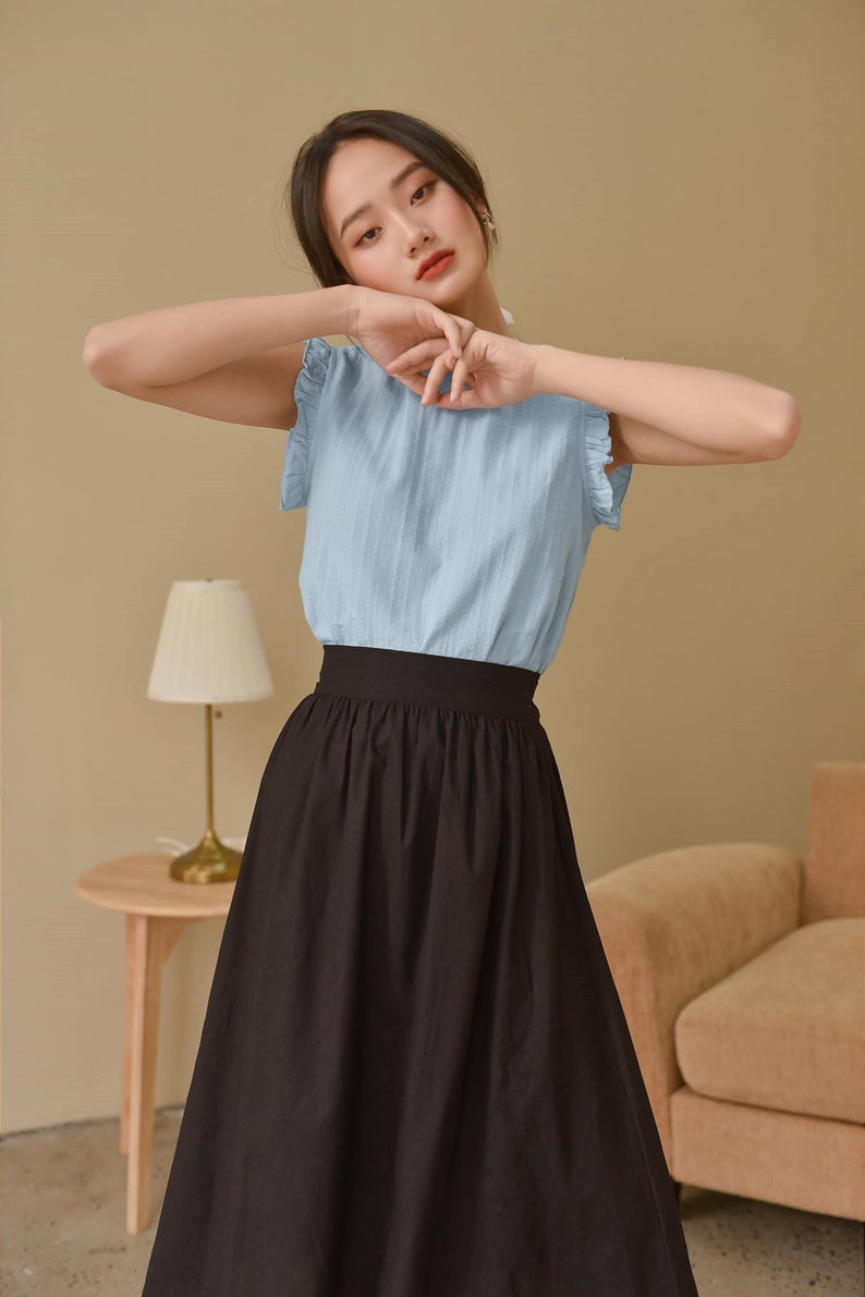 May include: A woman wearing a light blue, short-sleeved blouse with ruffled trim and a black, knee-length skirt.