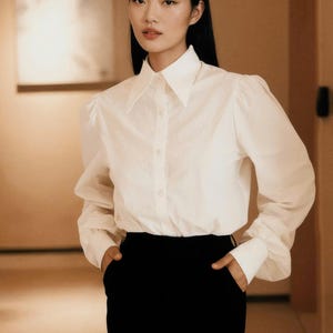 Puede incluir: Una camisa blanca abotonada con cuello ancho y mangas largas, combinada con una falda l&aacute;piz negra. La camisa tiene un dise&ntilde;o de manga ligeramente abullonada. El conjunto es de estilo cl&aacute;sico y elegante.
