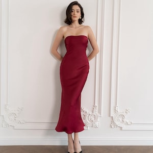 Puede incluir: Una mujer con un vestido de satén burdeos sin tirantes, con un corpiño ajustado y una falda fluida. Está de pie frente a una pared blanca con molduras ornamentadas.