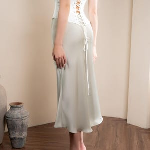 May include: A white corset top with a lace-up back and a light green satin skirt. The outfit is a formal style, with a high-waisted skirt and a strapless corset. The model is wearing high heels.