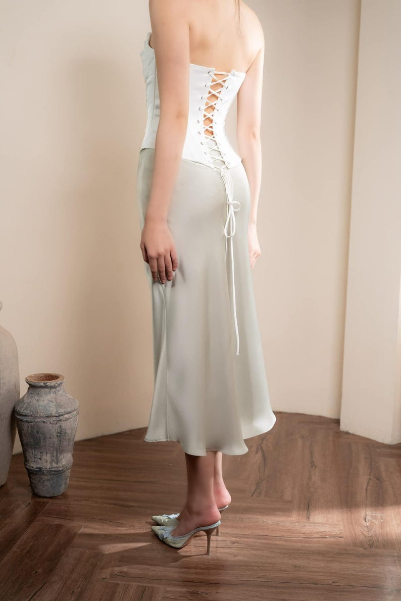 May include: A white corset top with a lace-up back and a light green satin skirt. The outfit is a formal style, with a high-waisted skirt and a strapless corset. The model is wearing high heels.
