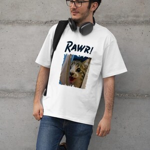 May include: White t-shirt with the word "Rawr!" in blue and a graphic of a cat with its mouth open. The cat graphic is framed in blue. The person is wearing glasses and jeans.