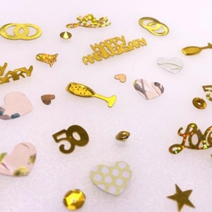 May include: A collection of gold-toned and patterned confetti pieces including the text Happy Anniversary, the number 50, interlocking rings, champagne flutes, stars, and hearts. The assorted shapes feature metallic, glittery, and printed finishes.