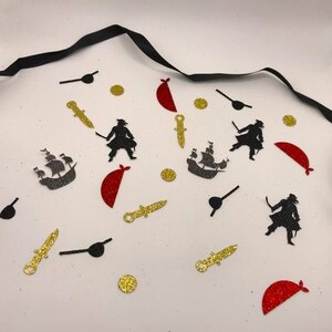 May include: A collection of black and gold glitter pirate themed confetti. The confetti includes images of pirates, ships, swords, eyepatches, and gold coins. There are also red glitter half-circles.