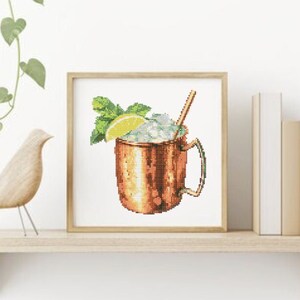 May include: A framed cross-stitch design of a copper mug filled with ice, a lime wedge, and a sprig of mint. A straw sticks out of the top of the mug.