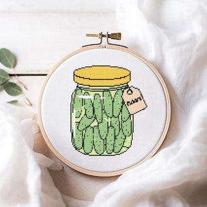 Op de afbeelding: Een witte borduurring met een kruissteekontwerp van een glazen pot gevuld met groene augurken. De pot heeft een geel deksel en een label met de tekst "Augurken".