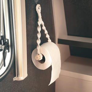 Könnte beinhalten: Eine Rolle weißes Toilettenpapier, aufgehängt an einem cremefarbenen Makramee-Halter. Der Halter ist mit einem kleinen Haken an einer dunklen Wand befestigt. Das Design ist einfach und funktional, passend für Bad oder Wohnmobil.