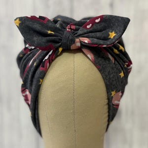 May include: A dark gray fabric headband with a large bow on top. The headband is patterned with pink lips, white clouds, and yellow stars. The bow is tied in the center and has two pointed ends.