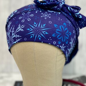 May include: A navy blue scrub cap with a snowflake pattern in shades of blue. The cap is tied in a bow at the back. The cap is designed to be worn in a medical setting.