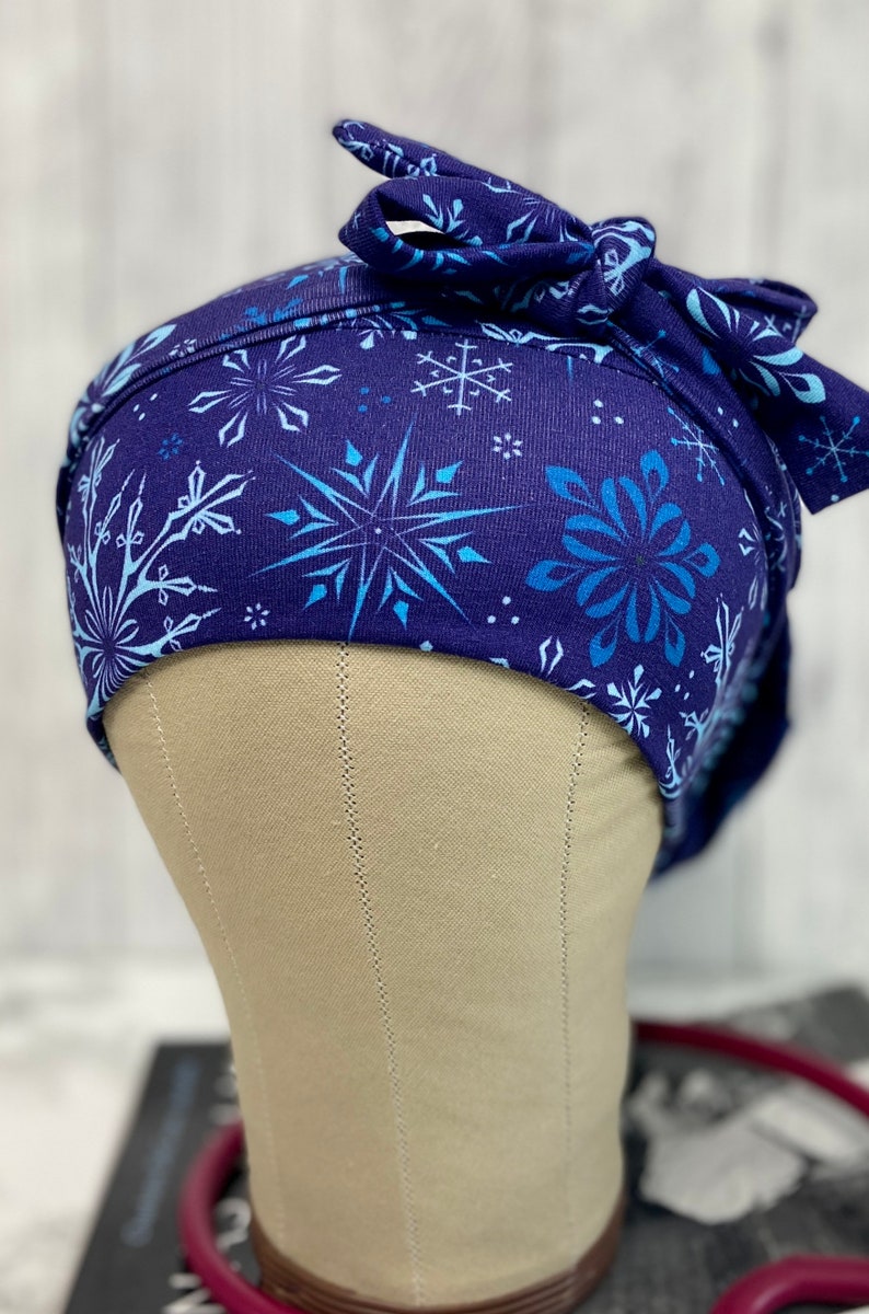 May include: A navy blue scrub cap with a snowflake pattern in shades of blue. The cap is tied in a bow at the back. The cap is designed to be worn in a medical setting.