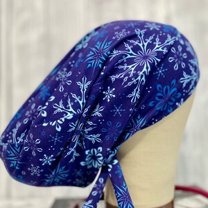 May include: A navy blue surgical scrub cap featuring a snowflake pattern in shades of light blue and white. The cap is tied at the back and is designed to cover the head and hair.
