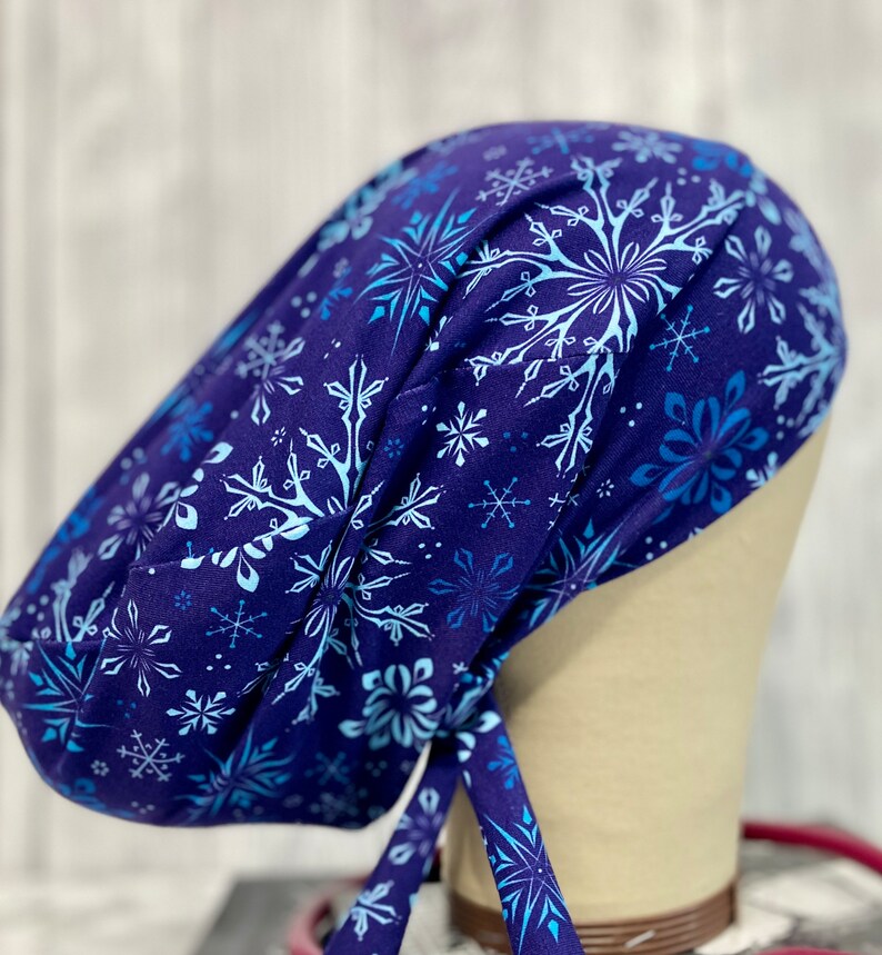 May include: A navy blue surgical scrub cap featuring a snowflake pattern in shades of light blue and white. The cap is tied at the back and is designed to cover the head and hair.