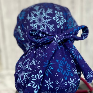May include: A navy blue scrub cap featuring a snowflake pattern in shades of light blue and white. The cap is tied in a bow at the back. The fabric is a soft, patterned material.