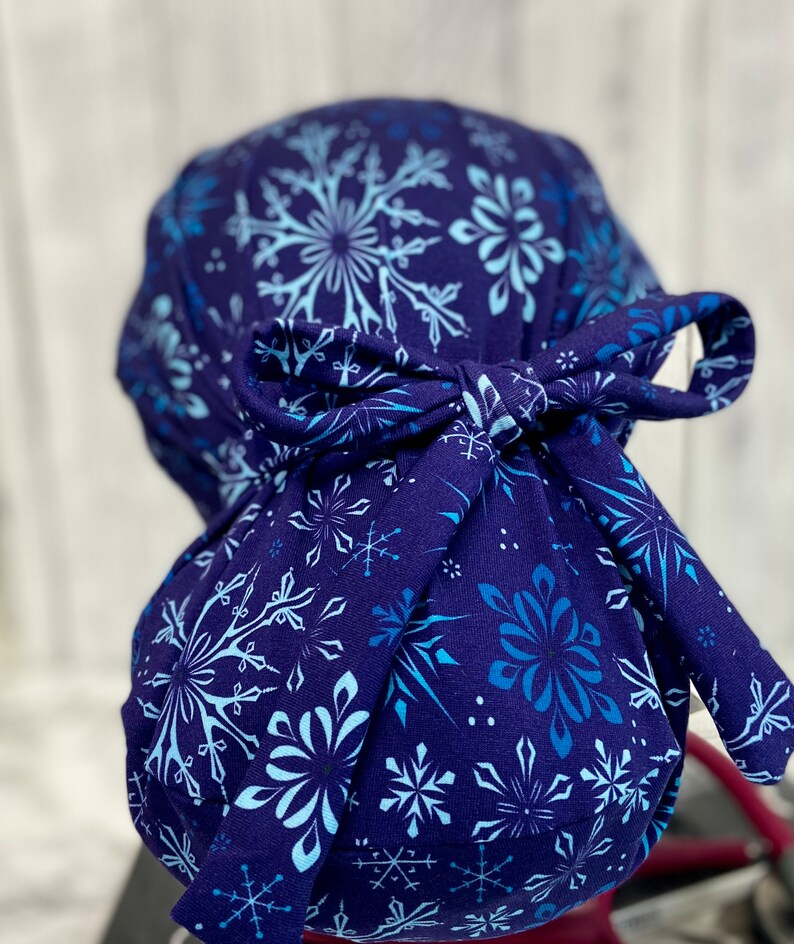 May include: A navy blue scrub cap featuring a snowflake pattern in shades of light blue and white. The cap is tied in a bow at the back. The fabric is a soft, patterned material.