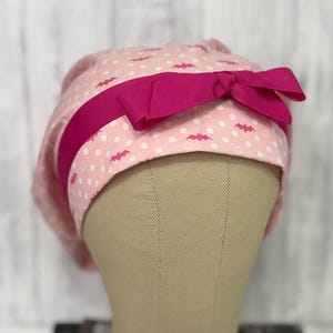 May include: A pink scrub cap with a pattern of white polka dots and pink bats. A bright pink ribbon is wrapped around the cap and tied into a bow. The cap is displayed on a mannequin head.