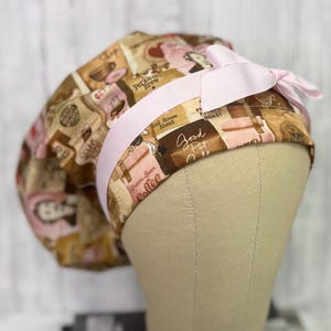 May include: A coffee-themed scrub cap with a light pink ribbon and bow. The cap features a repeating pattern of coffee-related imagery in shades of brown, pink, and cream, including text like "Dream Bean Coffee" and "Good Pot".