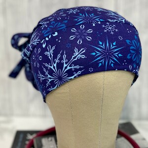 May include: A navy blue scrub cap featuring a repeating pattern of light blue and white snowflakes. The cap is tied at the back with matching fabric ties. The cap is displayed on a mannequin head.