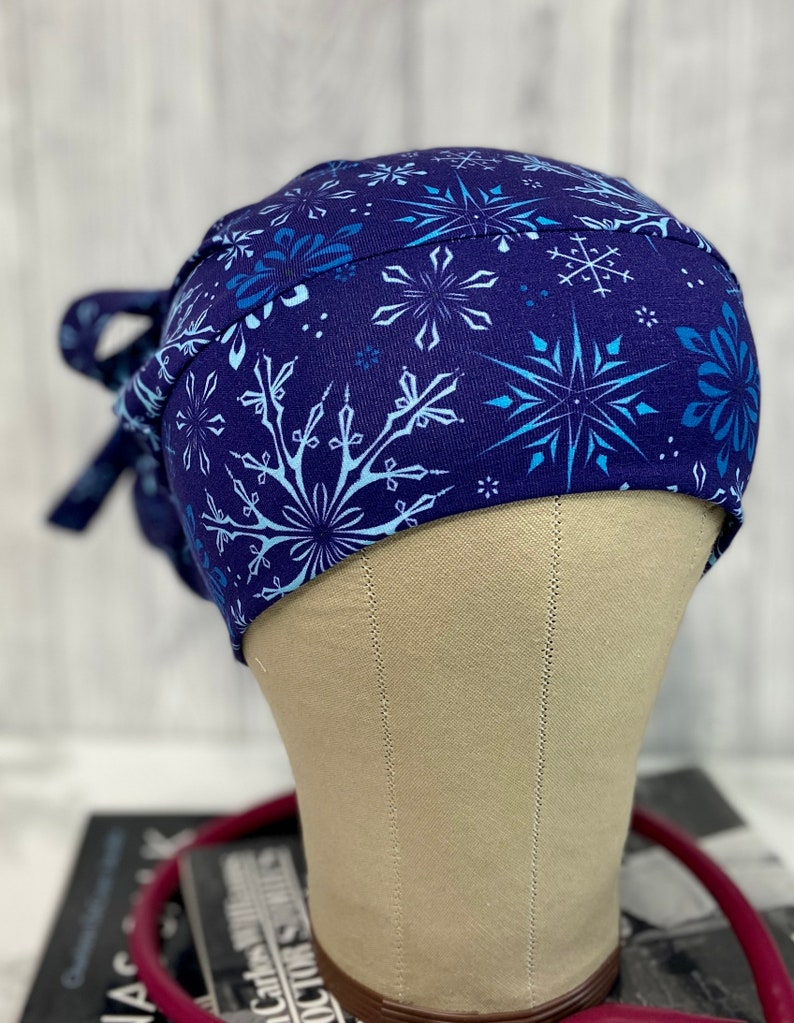 May include: A navy blue scrub cap featuring a repeating pattern of light blue and white snowflakes. The cap is tied at the back with matching fabric ties. The cap is displayed on a mannequin head.