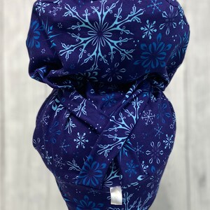 May include: A dark blue scrub cap with a repeating pattern of light blue snowflakes. The cap is made of soft fabric and has a gathered design at the back. The cap is designed to be worn in a medical setting.