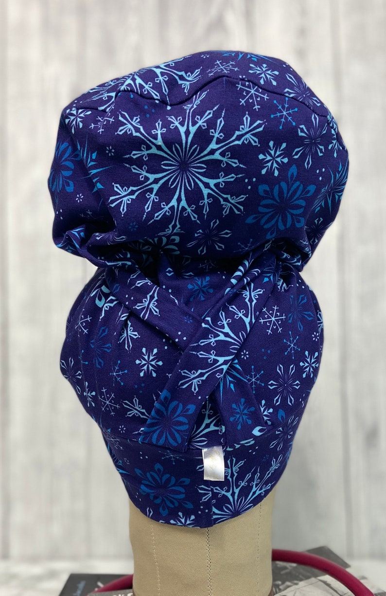 May include: A dark blue scrub cap with a repeating pattern of light blue snowflakes. The cap is made of soft fabric and has a gathered design at the back. The cap is designed to be worn in a medical setting.