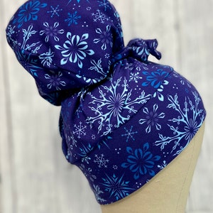May include: A dark blue scrub cap featuring a pattern of snowflakes in shades of blue and white. The cap is tied at the back and is designed to cover the hair. The fabric appears to be soft and comfortable.