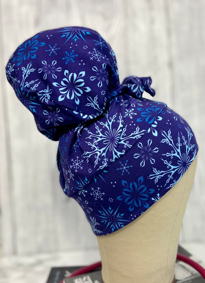 May include: A dark blue scrub cap featuring a pattern of snowflakes in shades of blue and white. The cap is tied at the back and is designed to cover the hair. The fabric appears to be soft and comfortable.