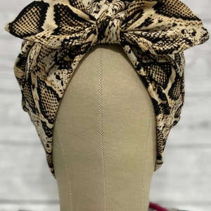 May include: A beige and black snakeskin print headband with a large bow on top. The headband is displayed on a mannequin head. The background is a blurred white wooden surface.