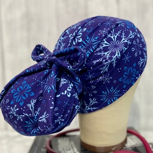 May include: A navy blue scrub cap with a snowflake pattern in shades of light blue and white. The cap is tied in a bow at the back. The cap is designed to be worn by medical professionals.
