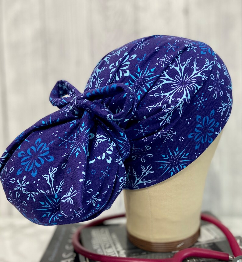 May include: A navy blue scrub cap with a snowflake pattern in shades of light blue and white. The cap is tied in a bow at the back. The cap is designed to be worn by medical professionals.