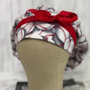 May include: A baseball-themed scrub cap featuring a pattern of white baseballs with red stitching on a gray background. A vibrant red ribbon and bow adorn the cap, adding a pop of color. The cap is designed for medical professionals or those in the healthcare field.
