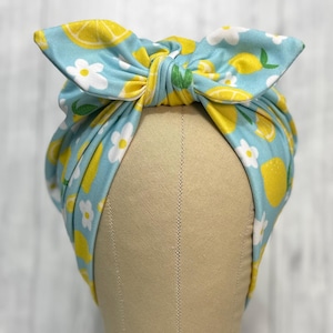 May include: A light blue fabric headband featuring a repeating pattern of yellow lemon slices, green leaves, and small white flowers. The headband is tied at the top into a large, prominent bow shape, creating a gathered and knotted appearance.
