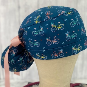 May include: A navy blue scrub cap featuring a repeating pattern of colorful bicycles with flower baskets. The cap has a pink ribbon bow on the side. The bicycles are in shades of yellow, pink, green, and orange.