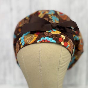 May include: A brown patterned scrub cap featuring a colorful turkey and floral design. The cap has a dark brown ribbon bow on top. The cap is displayed on a mannequin head.