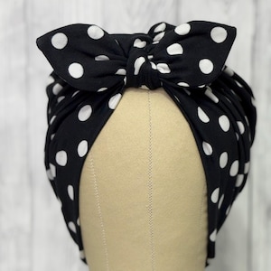 May include: A black head wrap featuring a white polka dot pattern and a large, knotted bow detail at the front. The fabric is gathered around the sides, creating a soft, draped appearance that covers the top and sides of the head form.