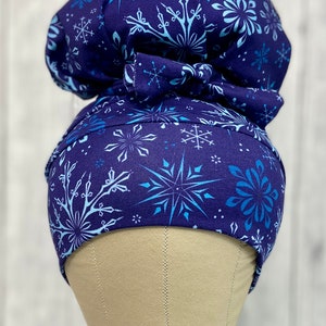 May include: A navy blue scrub cap with a snowflake pattern in shades of light blue and white. The cap features a bow detail and is displayed on a mannequin head. The overall theme is winter or a medical setting.