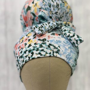 May include: A scrub cap featuring a floral pattern with light blue, pink, yellow, and dark green colors on a white background. The cap has a gathered back section and a fabric tie closure at the center, designed to secure the fit around the head.