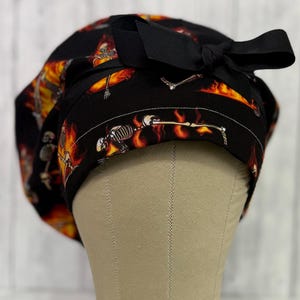 May include: Black surgical scrub cap with a repeating pattern of skeletons engulfed in flames. The cap has a black bow on the side. The design features orange, yellow, and white flames against a black background. The cap is made of fabric.