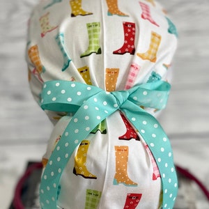Peut inclure: Une coiffe chirurgicale blanche ornée de bottes de pluie colorées, jaunes, vertes et rouges. Le bonnet est noué avec un ruban bleu clair à pois blancs. Conçu pour les professionnels de la santé.