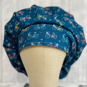 May include: A blue scrub hat with a pattern of colorful bicycles with baskets of flowers. The hat is on a  mannequin head.