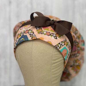 May include: A peach-colored scrub cap with a brown bow and trim. The cap features a floral and geometric pattern in shades of pink, blue, green, and yellow. The bow is centered on the cap's front, adding a decorative touch to the functional headwear.