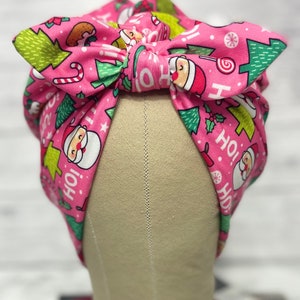 May include: Pink Christmas-themed fabric headband with a bow. The fabric features a repeating pattern of Santa Claus, Christmas trees, candy canes, and the words "Ho! Ho!"