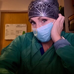 May include: A black and silver spiderweb patterned surgical cap. The cap is worn with a light blue surgical mask and a green scrub top. The image is a close-up of the person wearing the cap and mask.