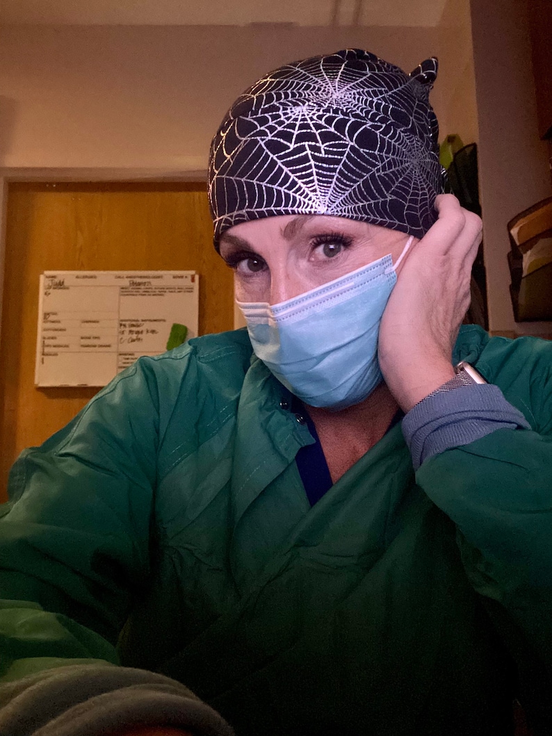 May include: A black and silver spiderweb patterned surgical cap. The cap is worn with a light blue surgical mask and a green scrub top. The image is a close-up of the person wearing the cap and mask.