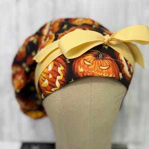 May include: A Halloween-themed scrub cap featuring a repeating pattern of carved jack-o'-lanterns on a black background. The cap has a yellow band and a matching bow. The cap is displayed on a mannequin head.
