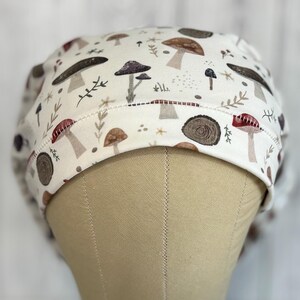 May include: A white scrub cap featuring a repeating pattern of various mushrooms in shades of brown, red, and gray, along with sprigs of greenery. The cap is designed to fit comfortably and securely, ideal for healthcare professionals or anyone needing hair protection.