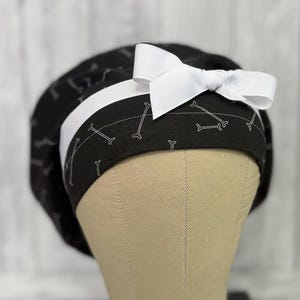 May include: A black surgical scrub cap with a white ribbon bow. The cap features a white bone pattern on a black background, with a white stripe. The cap is designed to be worn in a medical setting.