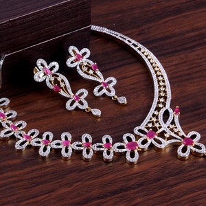 May include: A jewelry set consisting of a necklace and a pair of matching earrings. The pieces feature a floral-inspired design crafted with silver-toned metal, encrusted with small clear stones, and accented with teardrop-shaped pink-red stones.